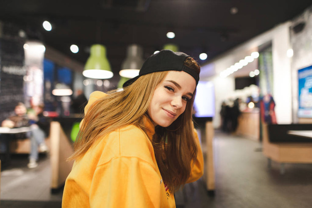 Glimlachend model in een cap en een oranje hoodie poses aan de camera binnenshuis, kijkt naar de camera en glimlach. Close-up portret - Foto, afbeelding