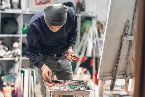 Professional artist in the studio at work, holding a brush in his hand and picking up the paint with a palette knife. Painting oil on canvas. Portrait of the painter at work. - Photo, Image
