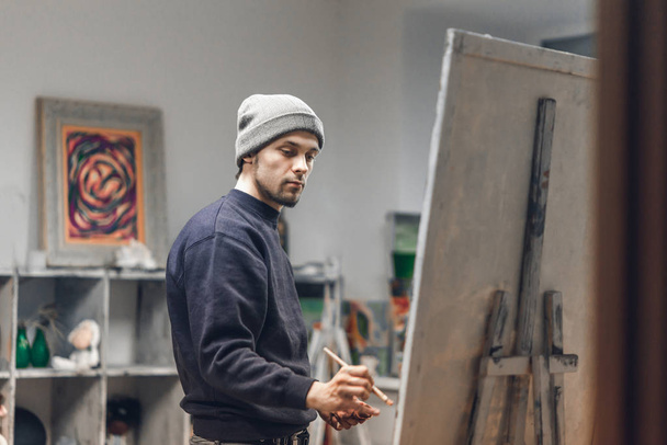 Busy man artist with a brush in his hand picks up an oil painting in a cozy studio. Talented young painter paints a painting on canvas. Painting Concept. Work as a hobby. - Fotoğraf, Görsel