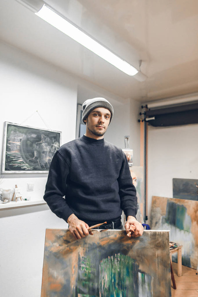 Portrait d'un artiste debout dans un studio avec une image et un pinceau dans les mains et regardant la caméra. Artiste garde sa photo et pose sur la caméra
 - Photo, image