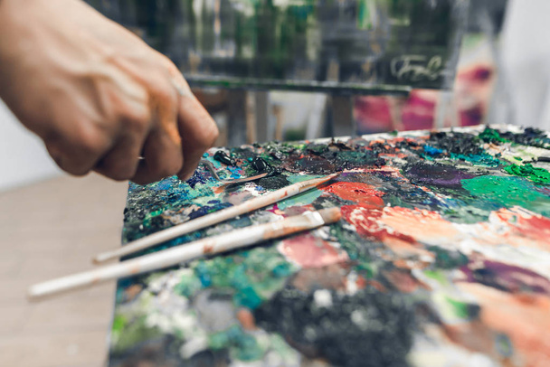 Brushes on a palette of oil paint, a hand of the artist with a knife palette takes a paint. Hands of the artist's husband are working on the picture. Background painting concept. Oil painting. - 写真・画像