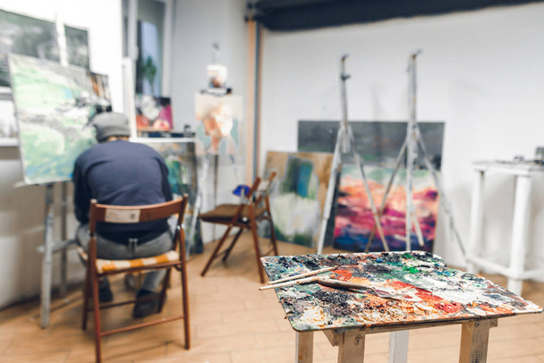 Palette and brush stand on the background of a painter sitting on a chair and paints a picture at the hostel in a cozy studio. Studio of painting. Painting workshop - Foto, afbeelding
