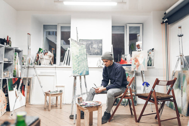 Young professional artist sits on a chair near an easel with a host in a cozy creative studio and paints an oil painting. Creative atmosphere in the home studio of painting. Interior - Fotoğraf, Görsel
