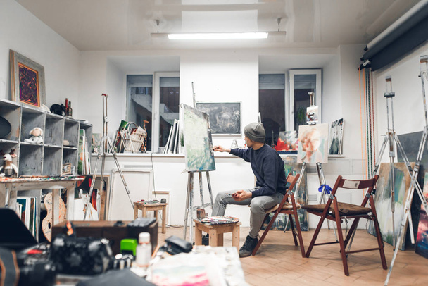画家は、油絵を描く居心地の良いスタジオと絵画の作品で椅子に座る。アーティストは、自宅のアトリエで動作します。趣味や仕事としての絵。インテリア アート スタジオ. - 写真・画像