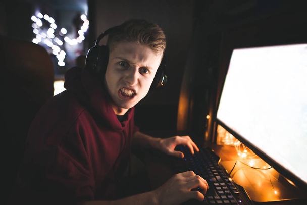 Nahaufnahme Porträt eines emotionalen Spielers mit Kopfhörern, spielt Videospiele auf einem Heimcomputer, blickt mit bösem Blick in eine Kamera. Ausdrucksstarker Gamer spielt Heimvideospiele. - Foto, Bild