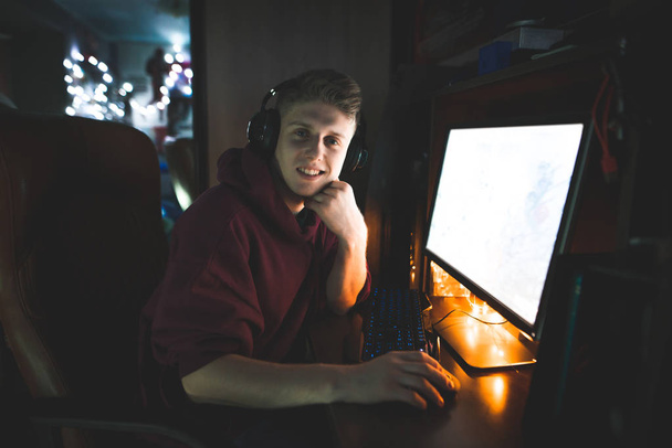 Porträt eines jungen attraktiven Mannes, der mit Kopfhörern am Computer sitzt, in die Kamera blickt und lächelt. Die glückliche Freiberuflerin arbeitet zu Hause am Computer. freiberufliches Konzept. - Foto, Bild
