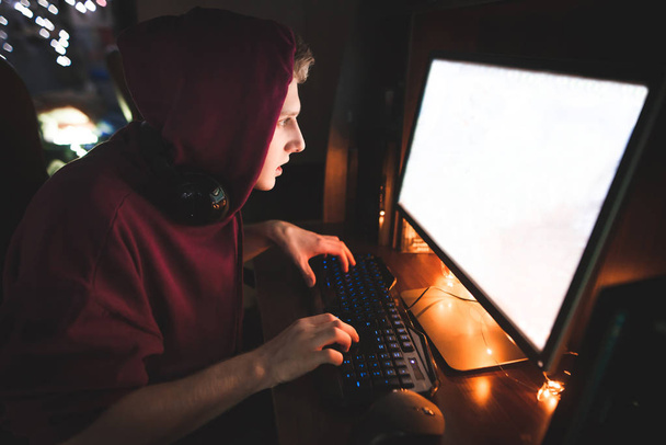 Концентрований підліток gamer в його капюшоном сидить в будинку в затишному залі і використовує комп'ютер. Gamer грає відео-ігор, фокусується на екрані комп'ютера. Концепція відеоігор. - Фото, зображення