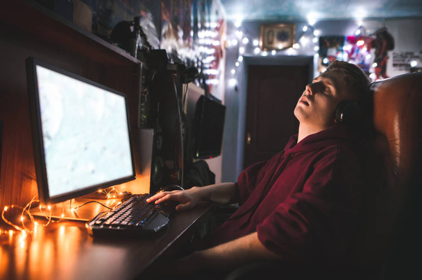 Mladý muž spí v křesle u počítače doma v útulné místnosti. Portrét hráč, který usnul z nudné počítačové hry. Gamer a Teenagery spí u stolu. - Fotografie, Obrázek
