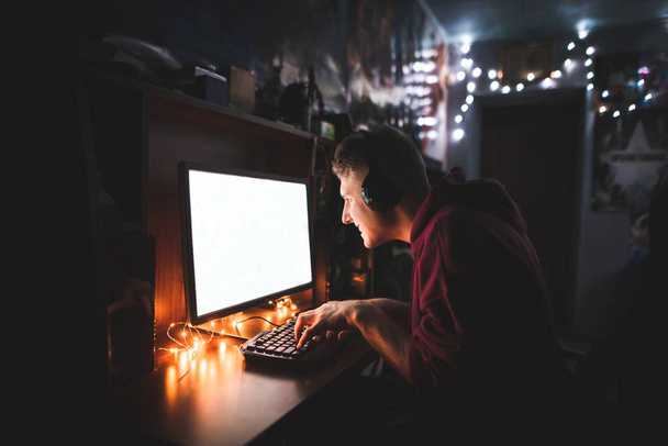 Портрет концентрированного молодого человека, играющего ночью в видеоигры на компьютере в уютной комнате дома. Игрок в наушники сидит дома в комнате и смотрит на экран компьютера
. - Фото, изображение