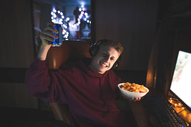 Portret van een vrolijke jongeman die gebruik maakt van een computer thuis 's nachts, houdt een pot van drankjes en platen met chips, kijkt naar de camera en glimlach. Portret van een tiener op een computer op een fast food. - Foto, afbeelding