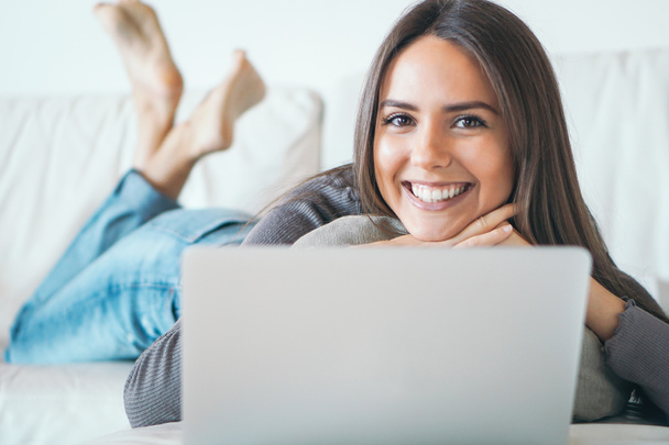ソファの上に横たわると自宅のラップトップ - 幸せな女の子カメラ微笑みながら彼女のコンピューターとオンラインでサーフィン - を使用して若い女性の人、技術、社会的なメディア ネットワークの概念 - 写真・画像