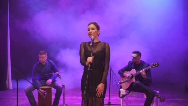 Music band performing on stage in different color light and smoke in background. Three musicians - girl singer, drummer and guitarist perform music during the show. - Footage, Video