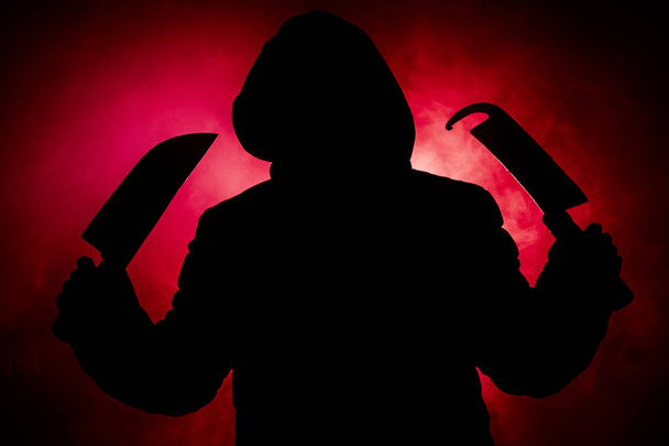 A dangerous hooded man standing in the dark and holding big knives. Face can not be seen. Committing a crime concept - Fotoğraf, Görsel
