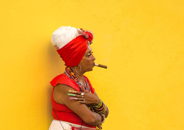 HAVANA-CUBA- DEC 4, 2018:  Woman with a cigar in her mouth with a yellow background letting tourists take photos of her for a few pesos in Havana street in Cuba.  It's a Cuban tradition. - Photo, image