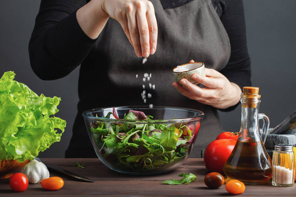 Woman chef in the kitchen preparing vegetable salad. Healthy Eating. Diet Concept. A Healthy Way Of Life. To Cook At Home. For Cooking. The girl sprinkles salt in a salad on a dark background. - Fotó, kép
