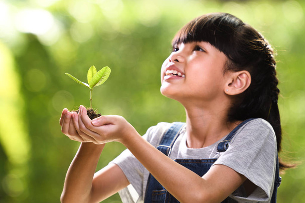 Uma menina segurando uma planta jovem em suas mãos com uma esperança de bom ambiente, foco seletivo na planta
 - Foto, Imagem