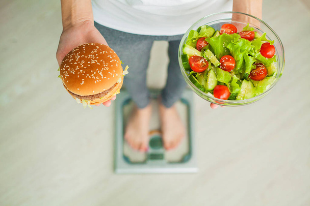 Δίαιτα. Γυναίκα μέτρηση σωματικού βάρους στη ζυγαριά κρατώντας Burger και σαλάτα. Τα γλυκά είναι ανθυγιεινά τρόφιμα παλιοπραγμάτων. Να κάνει δίαιτα, υγιεινή διατροφή, τον τρόπο ζωής. Απώλεια βάρους. Η παχυσαρκία. Το Top View - Φωτογραφία, εικόνα