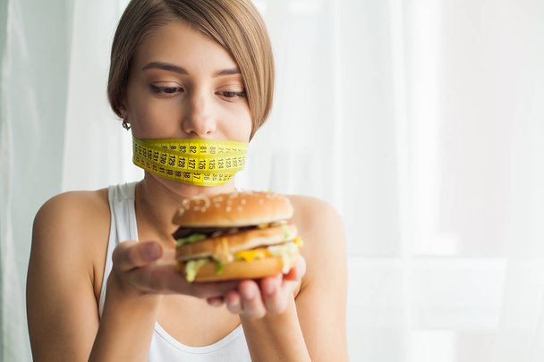 Diet concept and health. Emotional woman with measuring tapes around her mouth and unhealthy foodat her home - Photo, image