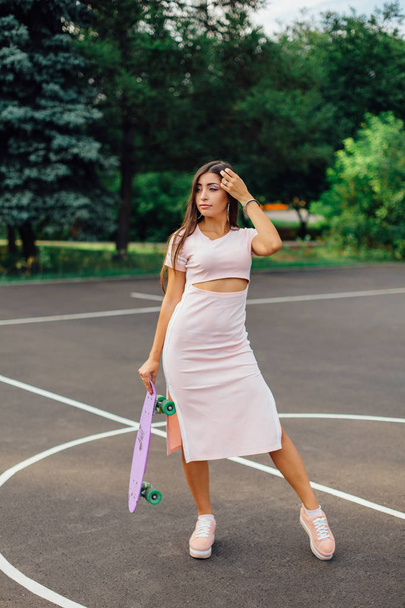 Portrait of a smiling charming brunette female holding in hand her skateboard on a basketball court. Happy woman with trendy look taking break during sunset. - Фото, изображение