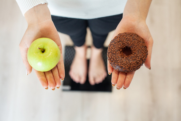 Kevyt. Nainen mittaamalla kehon paino punnitusasteikolla tilalla donitsi ja omena. Makeiset ovat epäterveellistä roskaruokaa. Ruokavalio, Terveellinen syöminen, Elämäntapa. Painonpudotus. Liikalihavuus
 - Valokuva, kuva