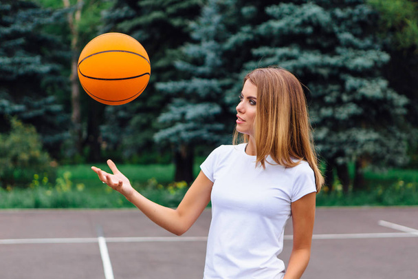 Beautiful young blonde girl dressed in white t-shirt, shorts and sneakers, plays with ball on a basketball court outdoors. Copy space. - Фото, изображение