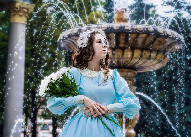 Portrait girl with a bouquet of white chrysanthemums in an elegant vintage dress stands by the fountain - Фото, изображение