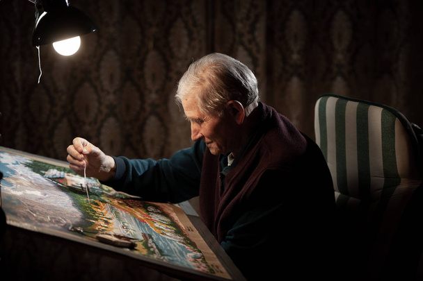 Elderly man woving a tapestry under bright light in living room on winter evening - Fotografie, Obrázek