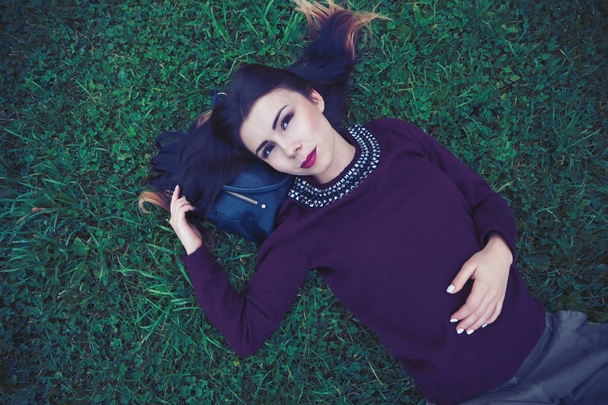 Beautiful young girl in stylish marsala sweater coat lying on the green grass with a smile. The model has red lipstick and bright make up - Valokuva, kuva