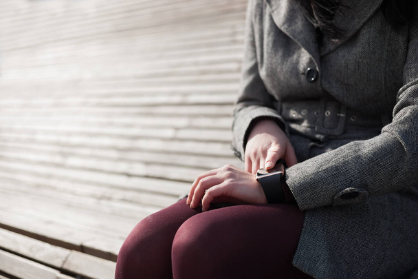 Female using her modern smart wrist watch sitting on the bench. This person is always connected to social media and internet. - Zdjęcie, obraz