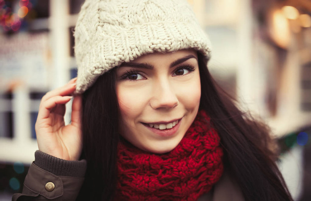 Happy young girl posing outdoor in warm winter clothes.Cold season fashion:massive knit red scarf and white hat on her head.Attractive friendly female model - Foto, Imagen