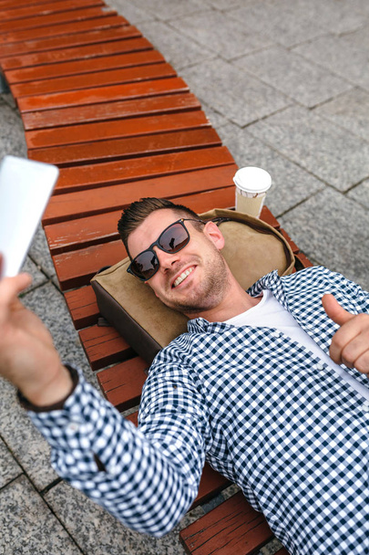 Мужчина лежит на скамейке в парке и делает селфи с мобильным телефоном.
 - Фото, изображение