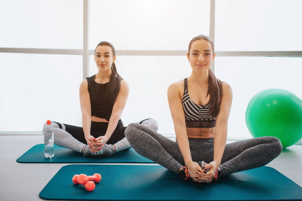 Two attractive and well-built young women sit on mat and stretch legs. They look on camera and smile. Models hold hands on feet. They sit in fitness room. - Фото, изображение