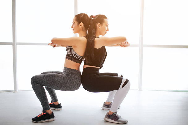 Two sloim young women exerising in fitness room. They keep hands crossed in chest area and touch each others back. Models sit in squad pose. - Foto, Imagen