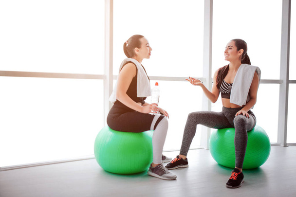 Tired but happy young women sit at fitballs and talk. They smile to each other. Models have white towels on their shoulders. They sit in fitness room. - Foto, Imagen