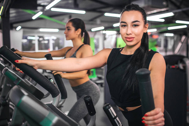 Nice young asian woman look on camera. She works on cross-trainer simulator. Another sexy young woman works at the same machine further. - Foto, imagen