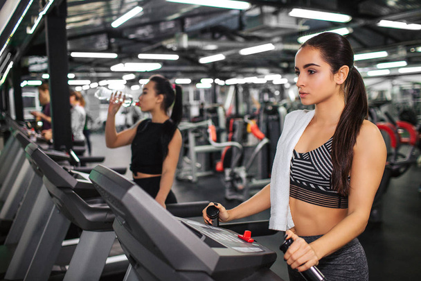 Serious and calm young woman look forward. She stand on running track and hold hands on it. Asian model walks on machine and drinks water. They work in gym. - Zdjęcie, obraz