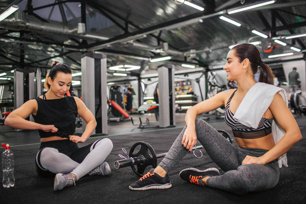 Picture of two young women sitting on floor in gym. Asian model tie knot on shirt and look down. Young european woman look at er and hold white towel on shoulder. - Photo, image
