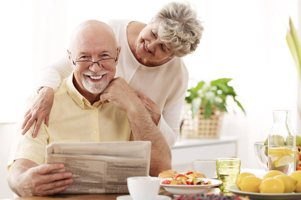 Happy senior couple hugging and smiling. Husband reading a newspaper - Fotó, kép
