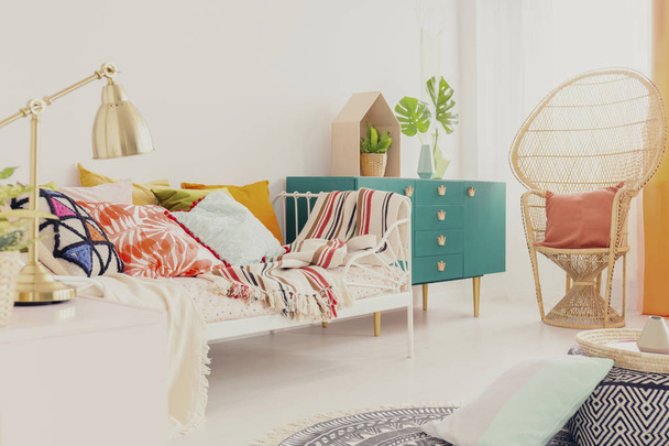 Χρυσό λυχνάρι στο nightstand στην κρεβατοκάμαρά του boho κοριτσιού με πολύχρωμα κλινοσκεπάσματα σε κρεβάτι, πράσινο ξύλινο ντουλάπι και παγώνι καρέκλα με μαξιλάρι - Φωτογραφία, εικόνα