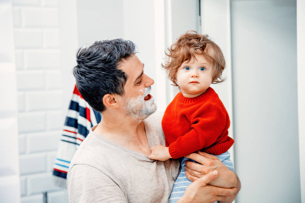 Vater mittleren Alters hält Kleinkind-Sonne und rasiert sich morgens im Badezimmer  - Foto, Bild