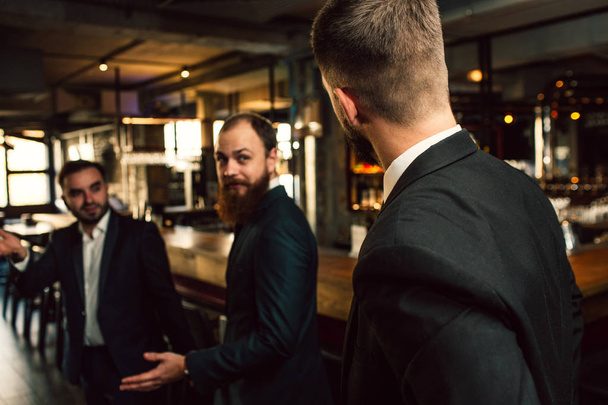 Three young men in suits lok at each other. One of them stand with back to camera. Office workers stand in pub. - Foto, Imagen
