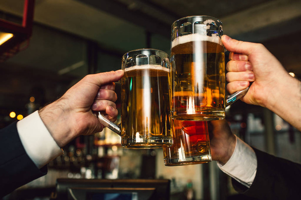 Picture of three mugs of beer in men hands. People wear suits. They are in bar. - Φωτογραφία, εικόνα