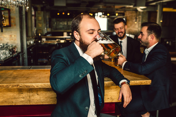 Young man in suit stand in bar and drink beer. Two other men stand behind him. They talk. - Φωτογραφία, εικόνα