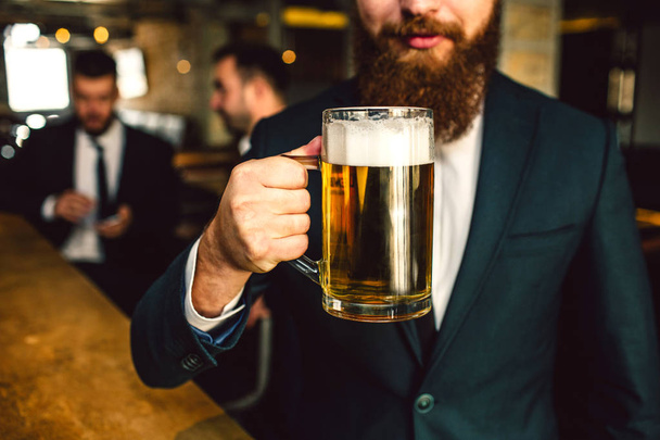 Cut view of bearded man in suit hold beer mug. Other two office workers sit behind. - 写真・画像