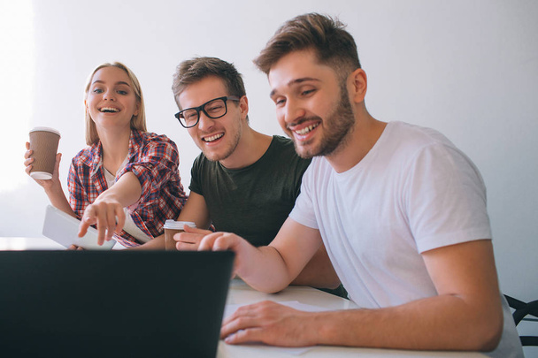Cheerful and positive young coworkers laughing. They sit at table in white room. People look at laptop screen. Young woman hold cup of coffee and point. - Foto, Bild
