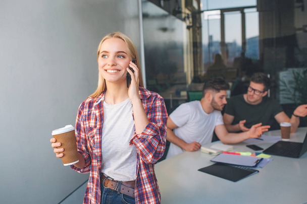 Cheerful young woman stand and talk on phone. She smiles. Model hold cup of coffee. Two young men sit behind her at table and work. - Foto, imagen