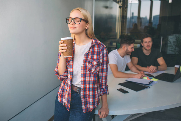Smart young blonde woman wear glasses. She stand and hold cup of coffee. She enjoyes. Two young men sit behind her and work. They talk between themselves. - Φωτογραφία, εικόνα