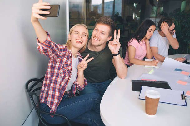 Happy young couple talking selfie. They sit together and look at phone camera. People show piece symbol. Another couple sit behind them and work at table. - Foto, Imagem