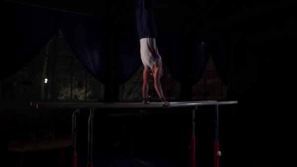 男性体操アクロバットがスローモーションで暗い部屋で平行棒に逆立ちを実行します。 - 映像、動画