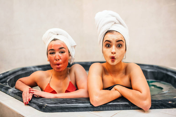 Amazed young women sit in jacuzzi and wonder. They look straight on camera. Women wear swimsuits and white towels. Models are amazed. They have beauty masks on face. - Photo, image
